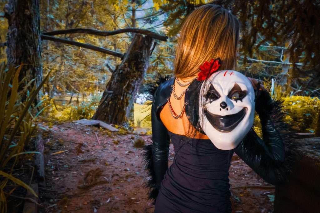 A woman in a black dress holding a clown mask in a forest, creating a mysterious and eerie scene.