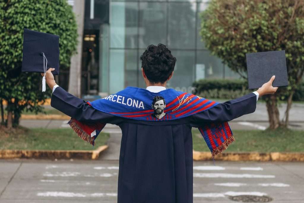Graduate in cap and gown holding Barcelona scarf outdoors.