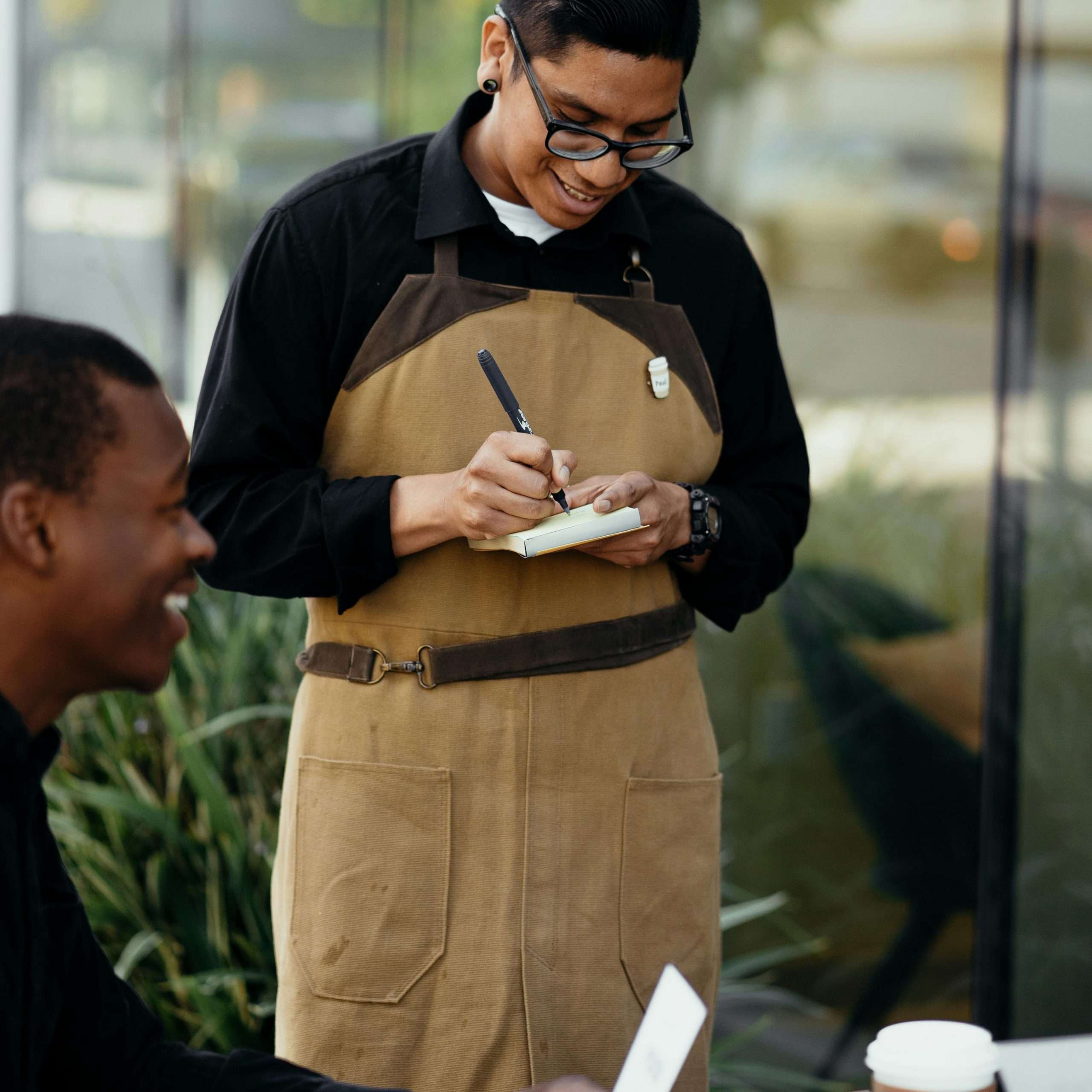 A smiling waiter takes an order from customers seated outside a café.