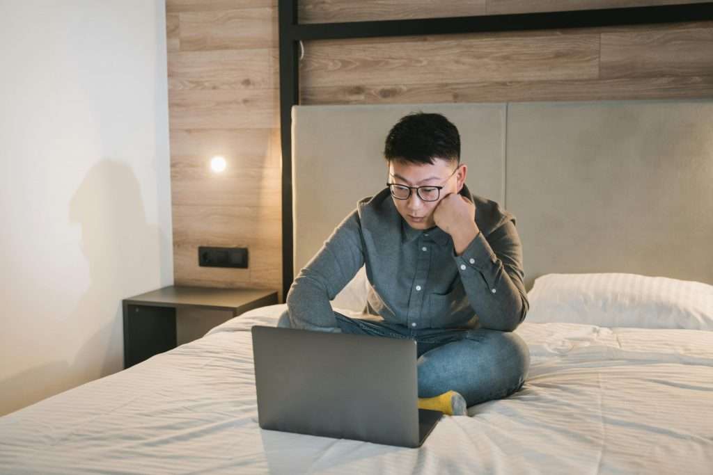 Asian man with glasses sitting on a bed, focused on his laptop, working remotely.
